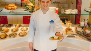 A chef in a white uniform holding a plate with a dessert, standing in front of a buffet with various food items and a floral centerpiece. - HOTEL POUSADA DA NEVE (Nova Petropolis)