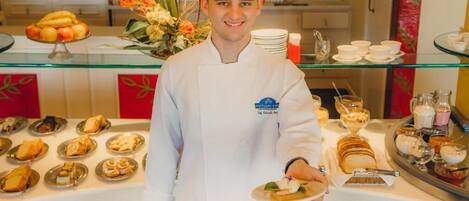 Un chef con uniforme blanco sostiene un plato con postre, frente a un bufé con diversas opciones de comida y un centro de mesa floral.