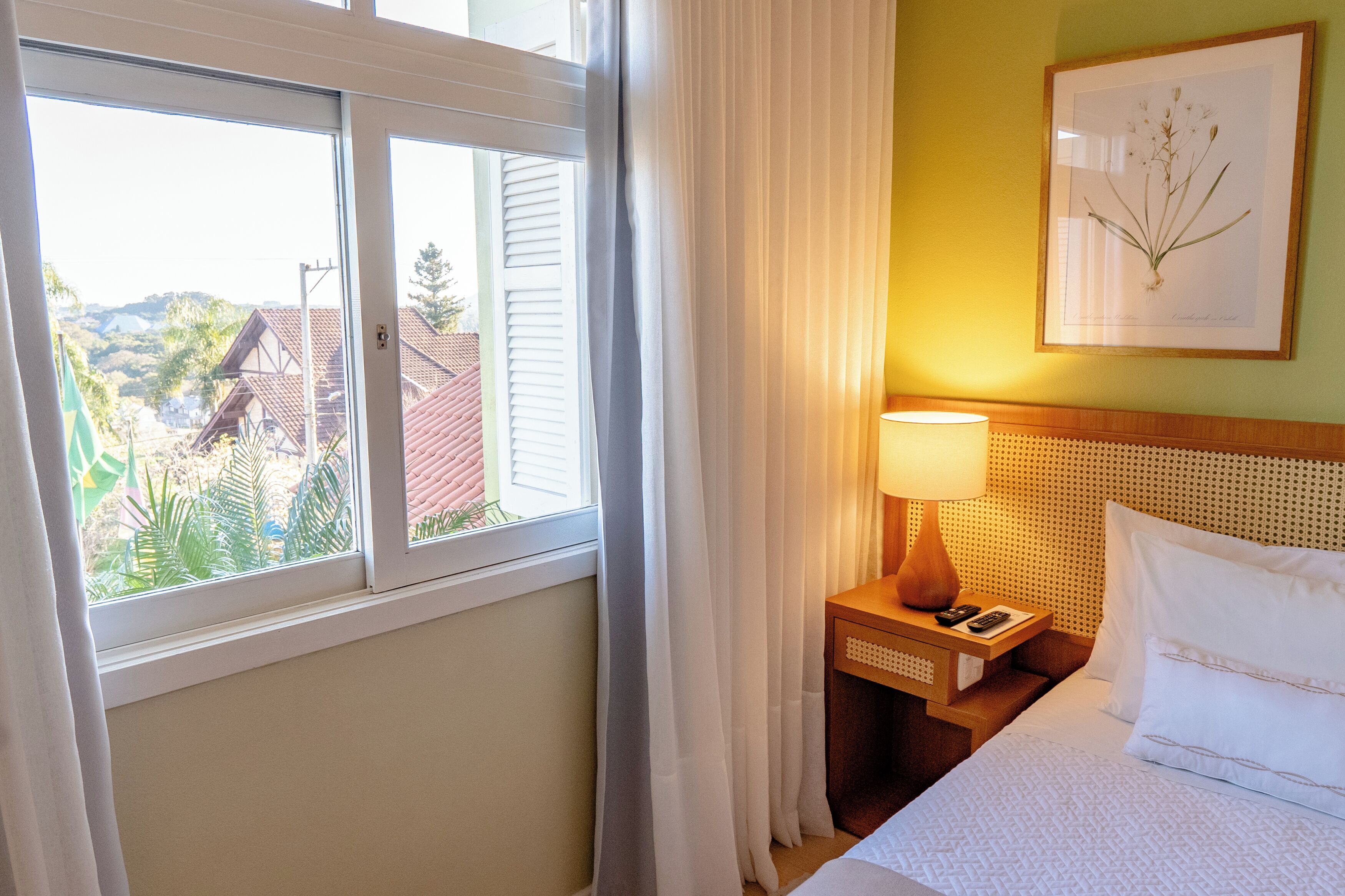A hotel room with a bed, a nightstand, a lamp, a framed botanical print, and a window with curtains.