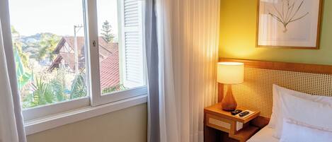 A hotel room with a bed, a nightstand, a lamp, a framed botanical print, and a window with curtains.