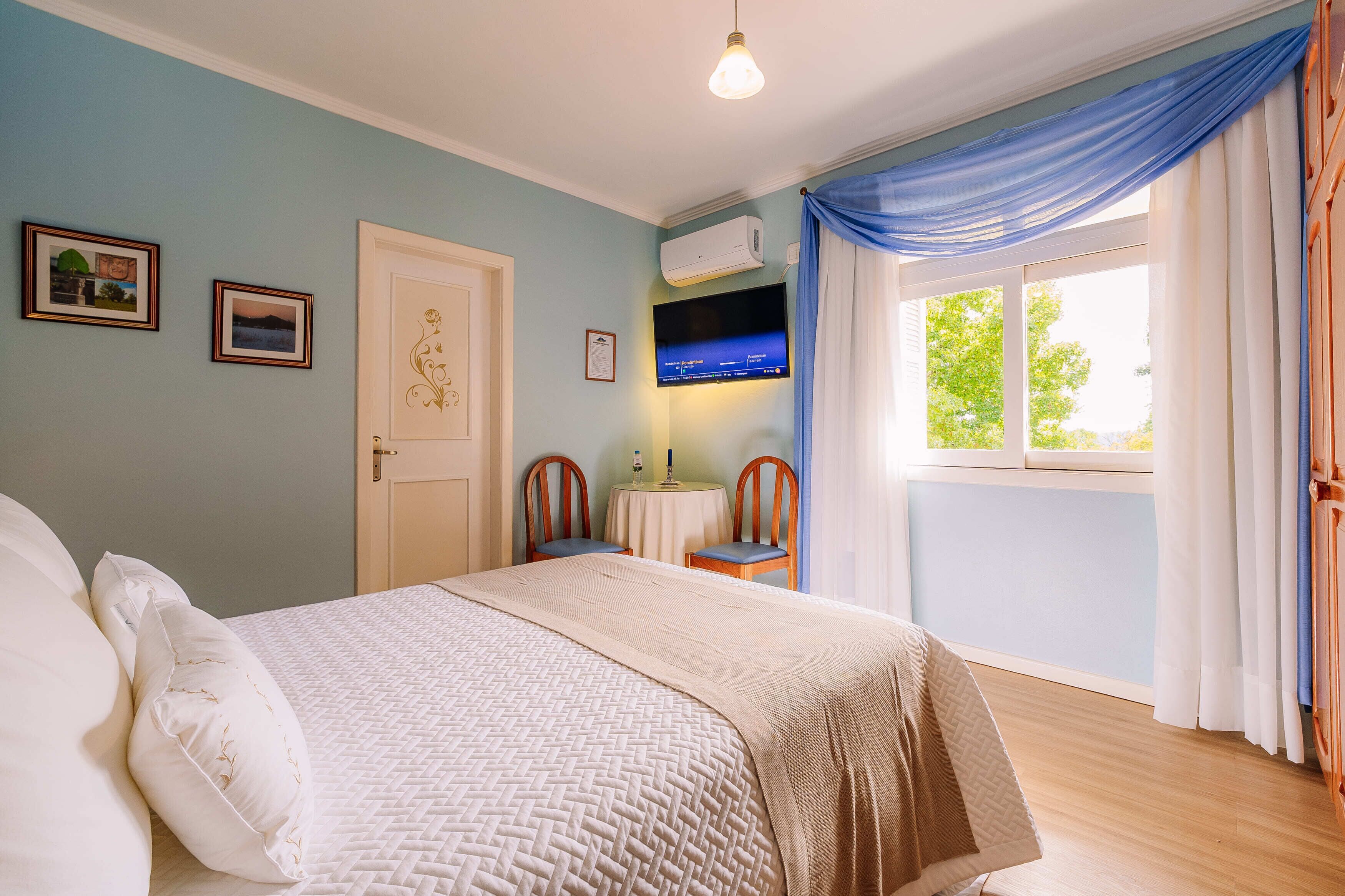 A bedroom with a bed, a TV, a window with curtains, and a door.