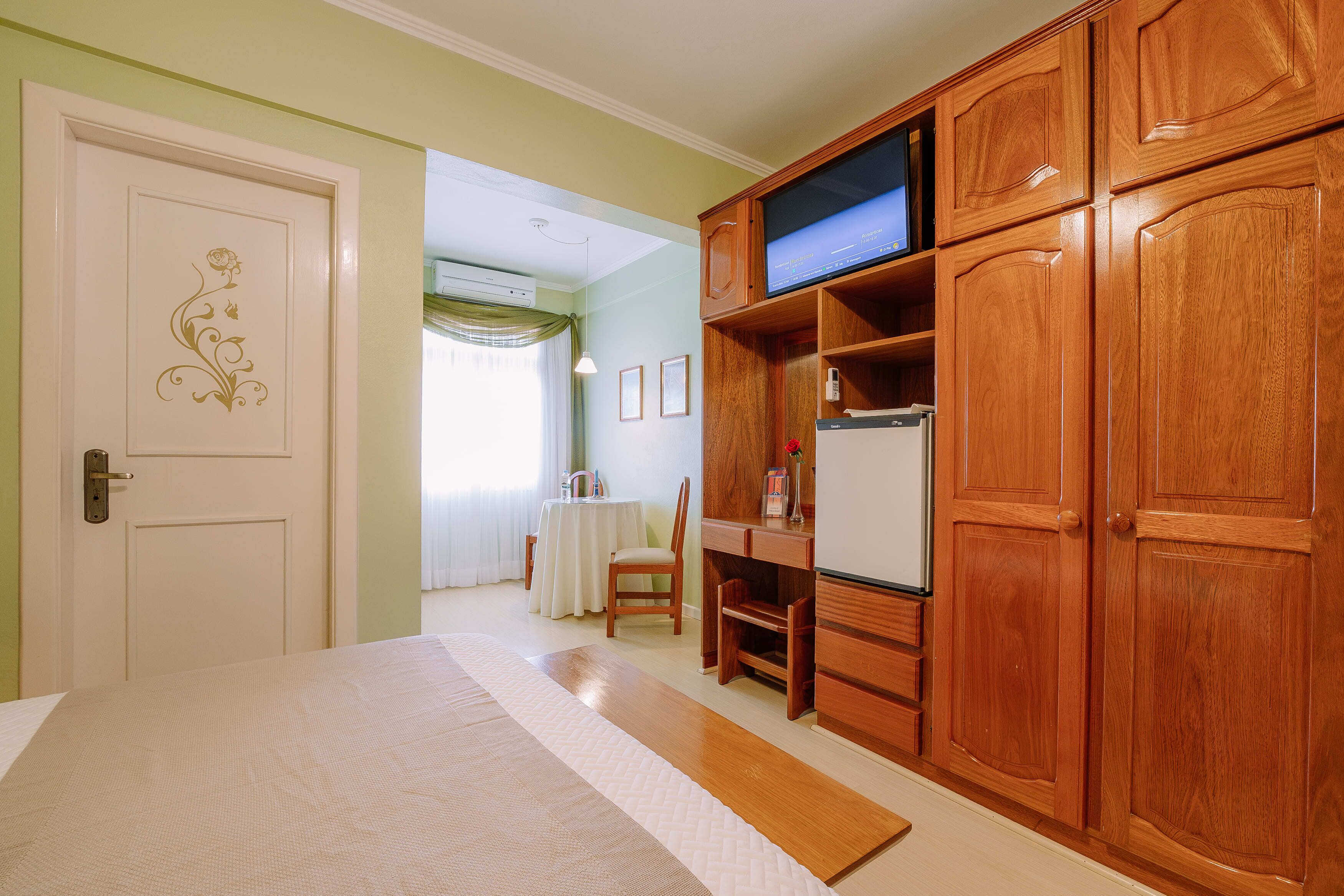 A bedroom with a bed, wooden wardrobe, a television, and a small table with a chair.
