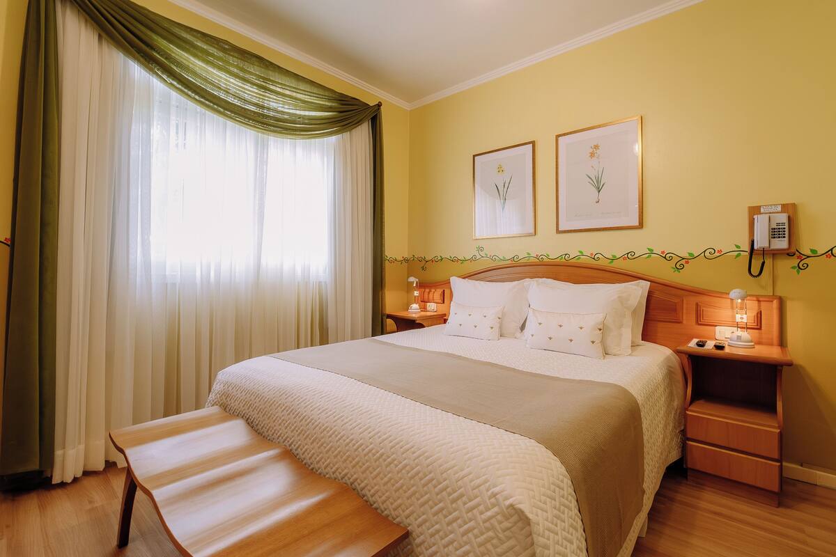 A hotel room with a large bed, a bedside table with a lamp, a bench, and a window with curtains.