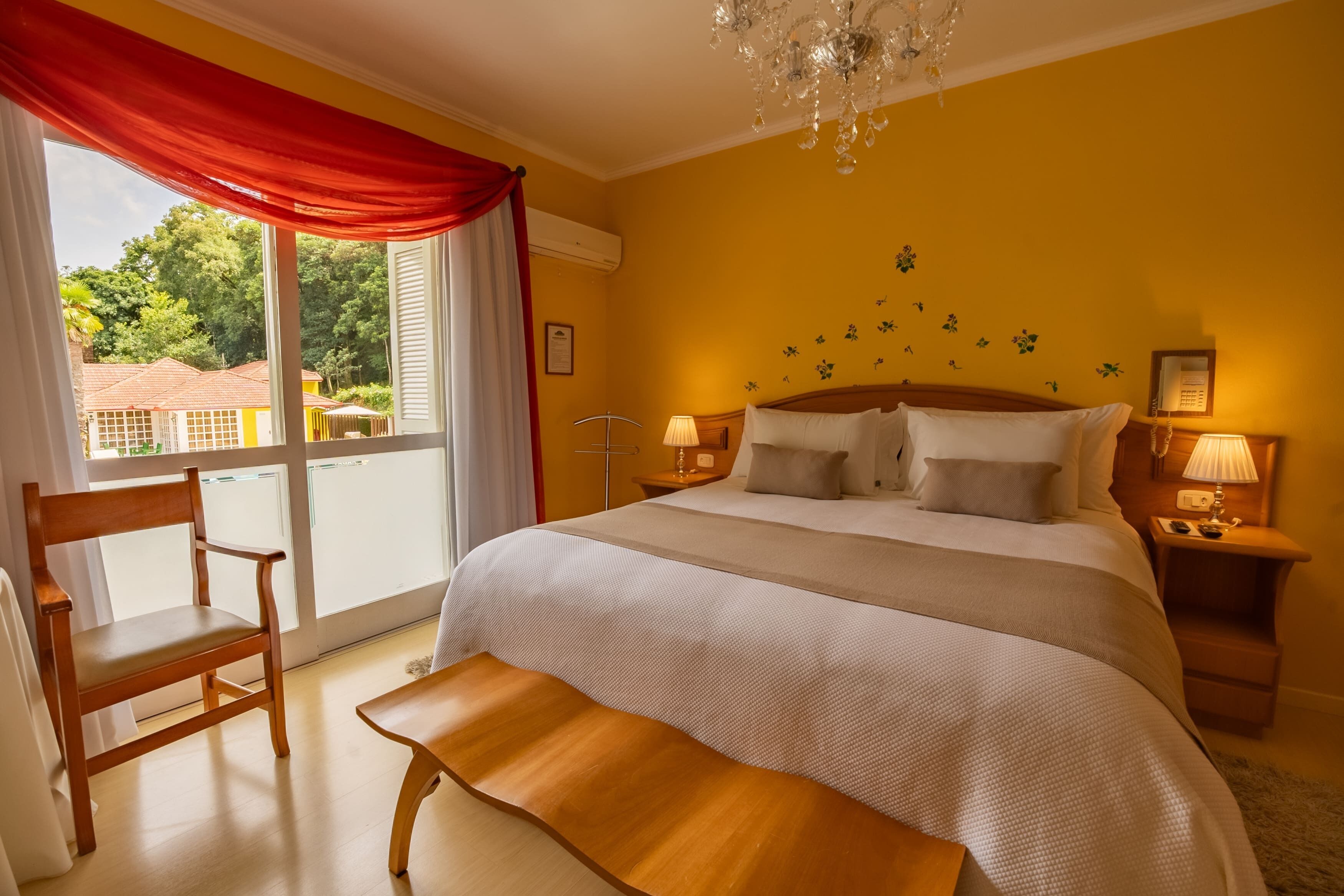 Habitación de hotel con una cama grande, una silla de madera, un banco, una araña y vistas al exterior.