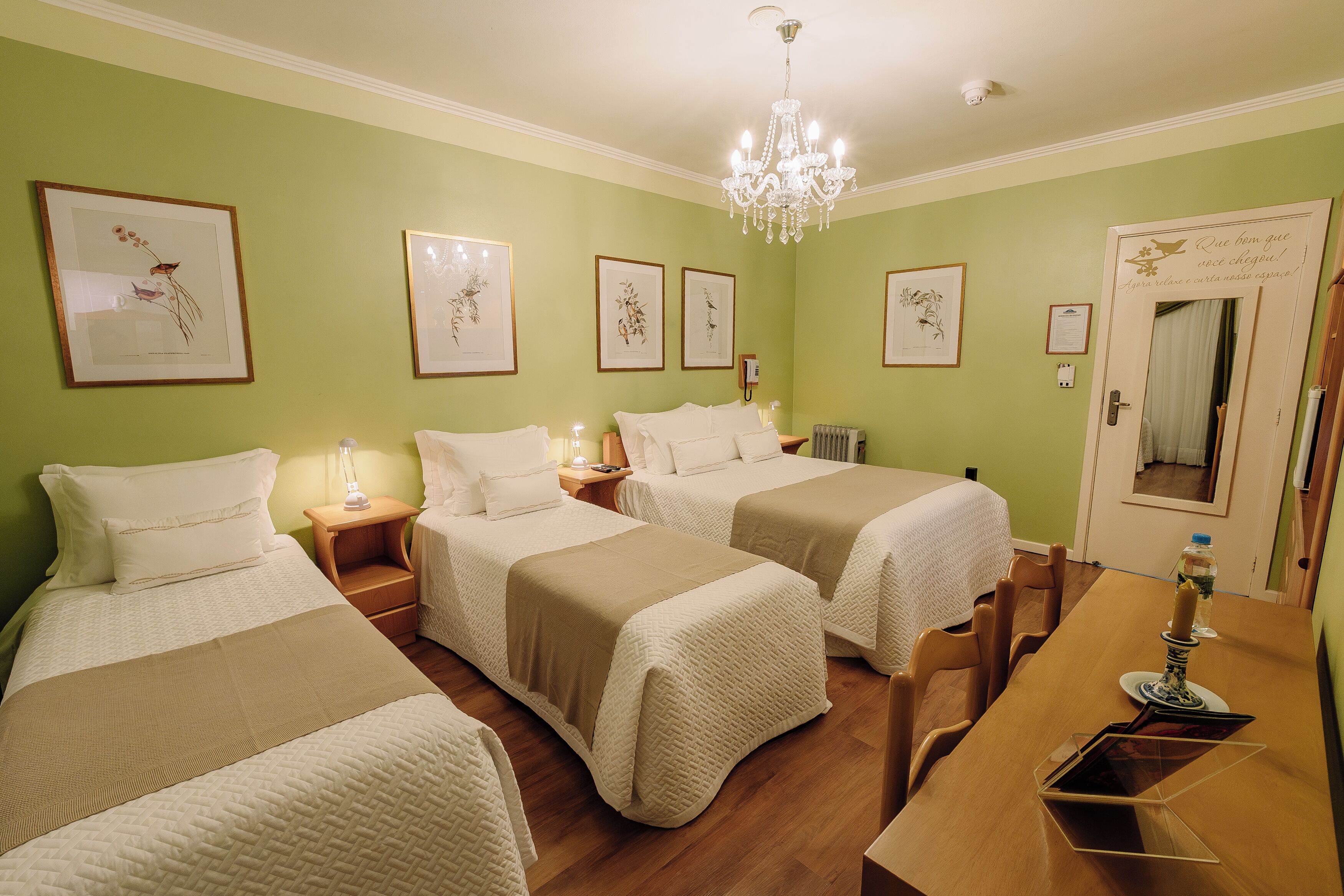 Habitación de hotel con tres camas, un candelabro, reproducciones botánicas enmarcadas en la pared y una mesa de comedor de madera.