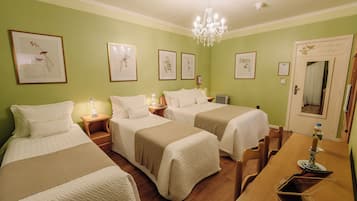 Habitación de hotel con tres camas, un candelabro, reproducciones botánicas enmarcadas en la pared y una mesa de comedor de madera.
