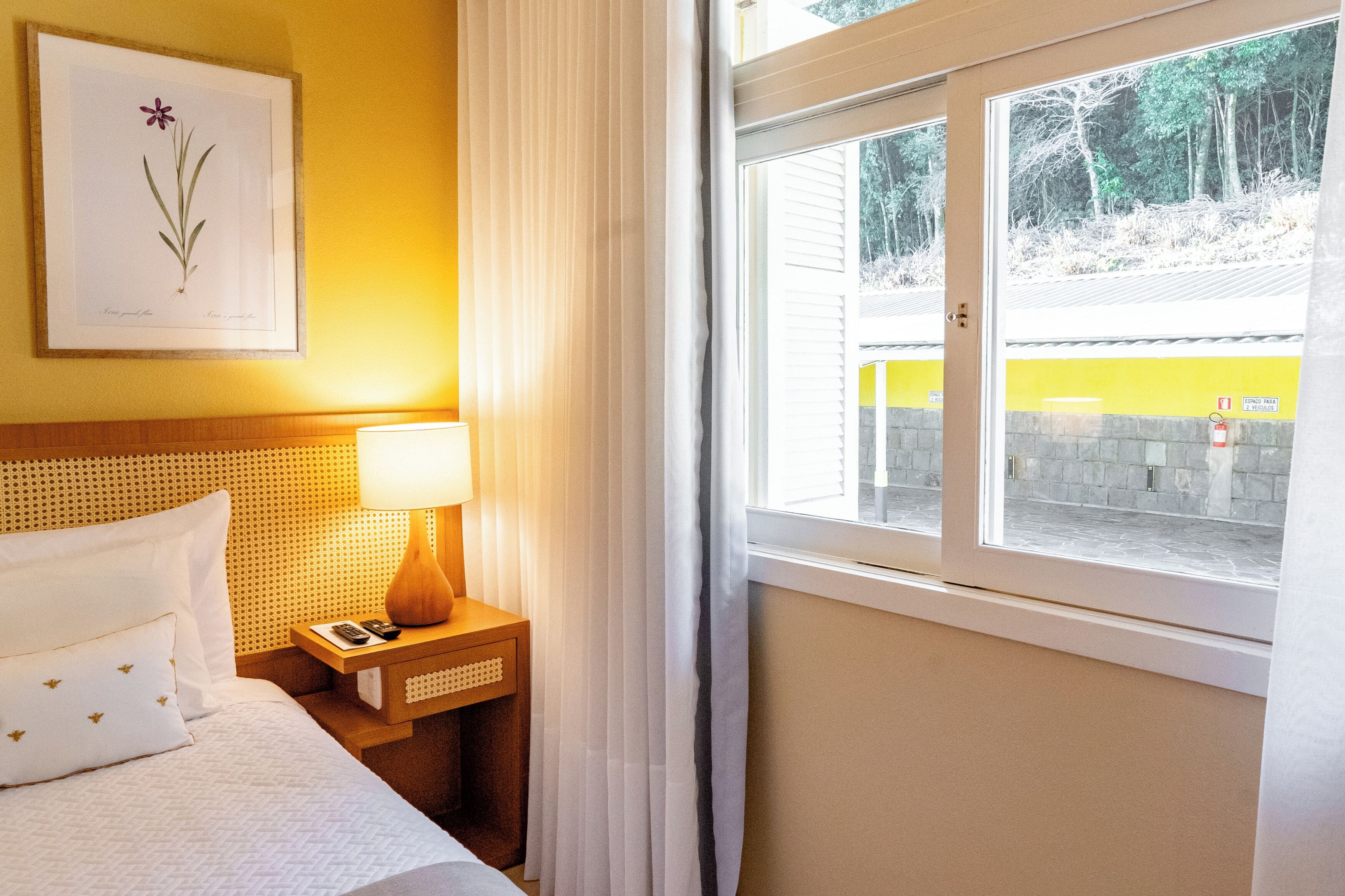 A hotel room with a bed, a nightstand with a lamp, a framed botanical print, and a window with a view of a yellow building and trees.