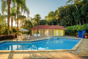 A swimming pool with a tiled edge, surrounded by a stone patio. There is a small building with a red roof and a yellow wall, and a white umbrella. - HOTEL POUSADA DA NEVE (Nova Petropolis)