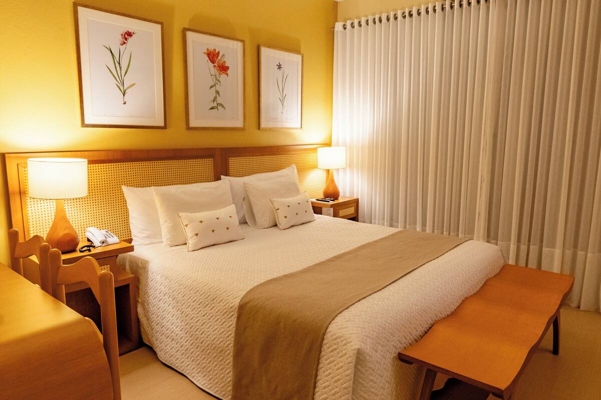 A hotel room with a large bed, two bedside tables with lamps, a wooden bench, and three framed floral artworks on the wall.