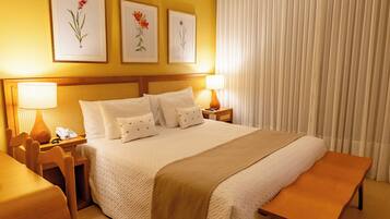 Une chambre d’hôtel avec un grand lit, deux tables de chevet avec des lampes, un banc en bois et trois œuvres d’art florales encadrées accrochées au mur.