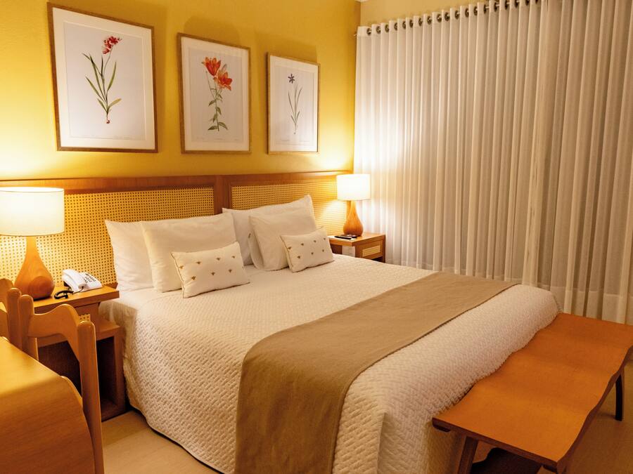 Une chambre d’hôtel avec un grand lit, deux tables de chevet avec des lampes, un banc en bois et trois œuvres d’art florales encadrées accrochées au mur.