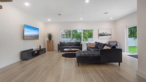A modern living room with a sectional sofa, a coffee table, and a flat-screen TV on a stand. There are large windows providing natural light and views of greenery outside. - Luxury 4BR Retreat w/ Heated Pool + Free Car Rental – Near Miami & Beaches (Biscayne Park)