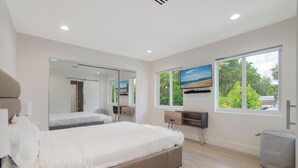 A modern bedroom with a large bed, a mirror, a TV, and a window with a view of trees and a beach. - Luxury 4BR Retreat w/ Heated Pool + Free Car Rental – Near Miami & Beaches (Biscayne Park)