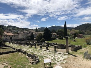 An ancient ruin with stone columns and arches, surrounded by a grassy area and a village in the background.