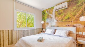 A bedroom with a large bed, a window with a view of trees, a wall-mounted air conditioner, and a wooden headboard. - PF42 Linda Casa 4 Quartos- Villa Firenze (Mata de São João)