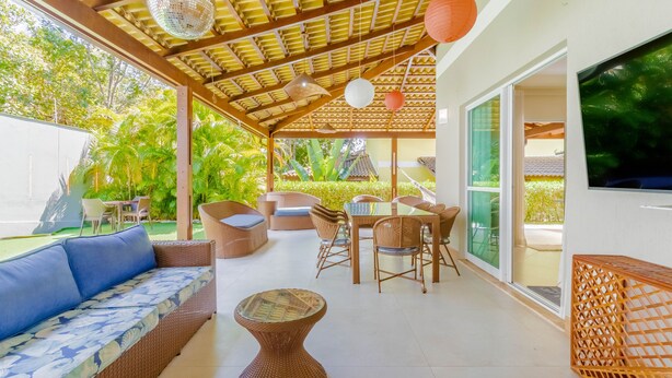 A covered outdoor seating area with wicker furniture, a glass-top table, and a TV screen on the wall. - PF42 Linda Casa 4 Quartos- Villa Firenze (Mata de São João)
