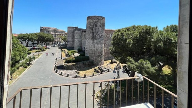 A view of a historic castle with a central tower, surrounded by greenery and a paved area with people walking. - Suite Ursino B&B (Catania)