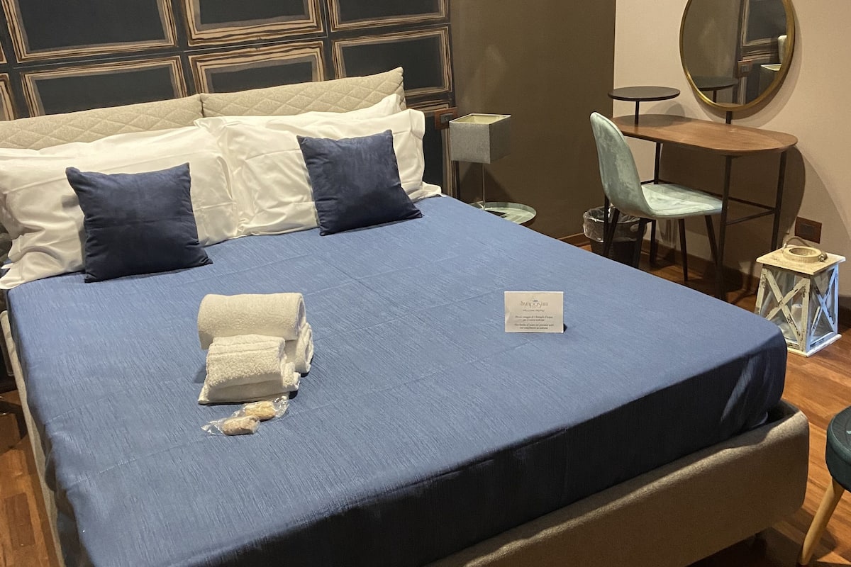 Cama bem arrumada com colcha e travesseiros azuis, uma pequena mesa de cabeceira com abajur, uma cadeira e um espelho redondo na parede.