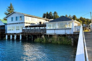 Una propiedad frente al agua con un muelle de madera que se adentra en el agua, una casa con balcón y un edificio aparte con grandes ventanales.