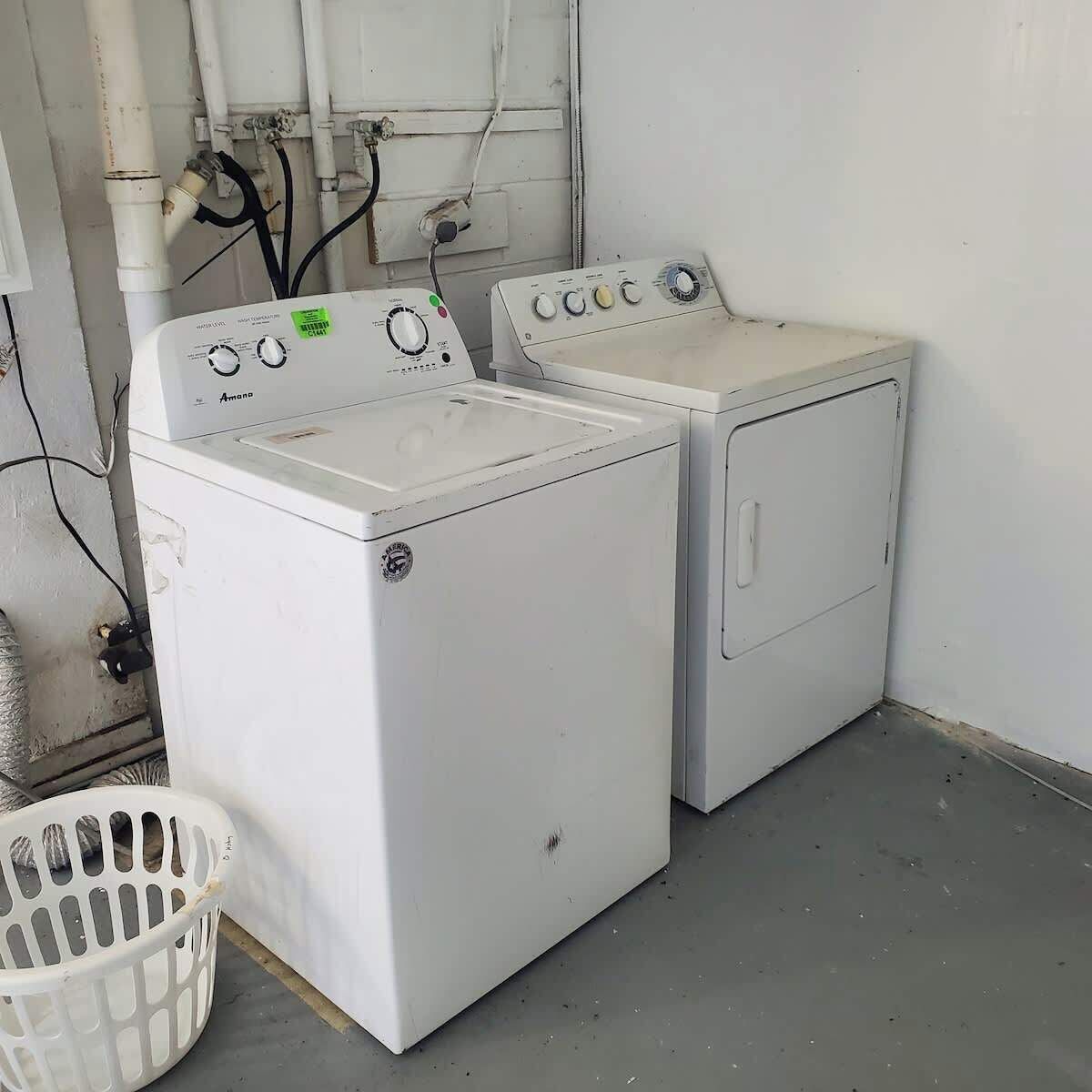 A laundry room with two white front-loading washing machines, a white plastic laundry basket, and visible plumbing.