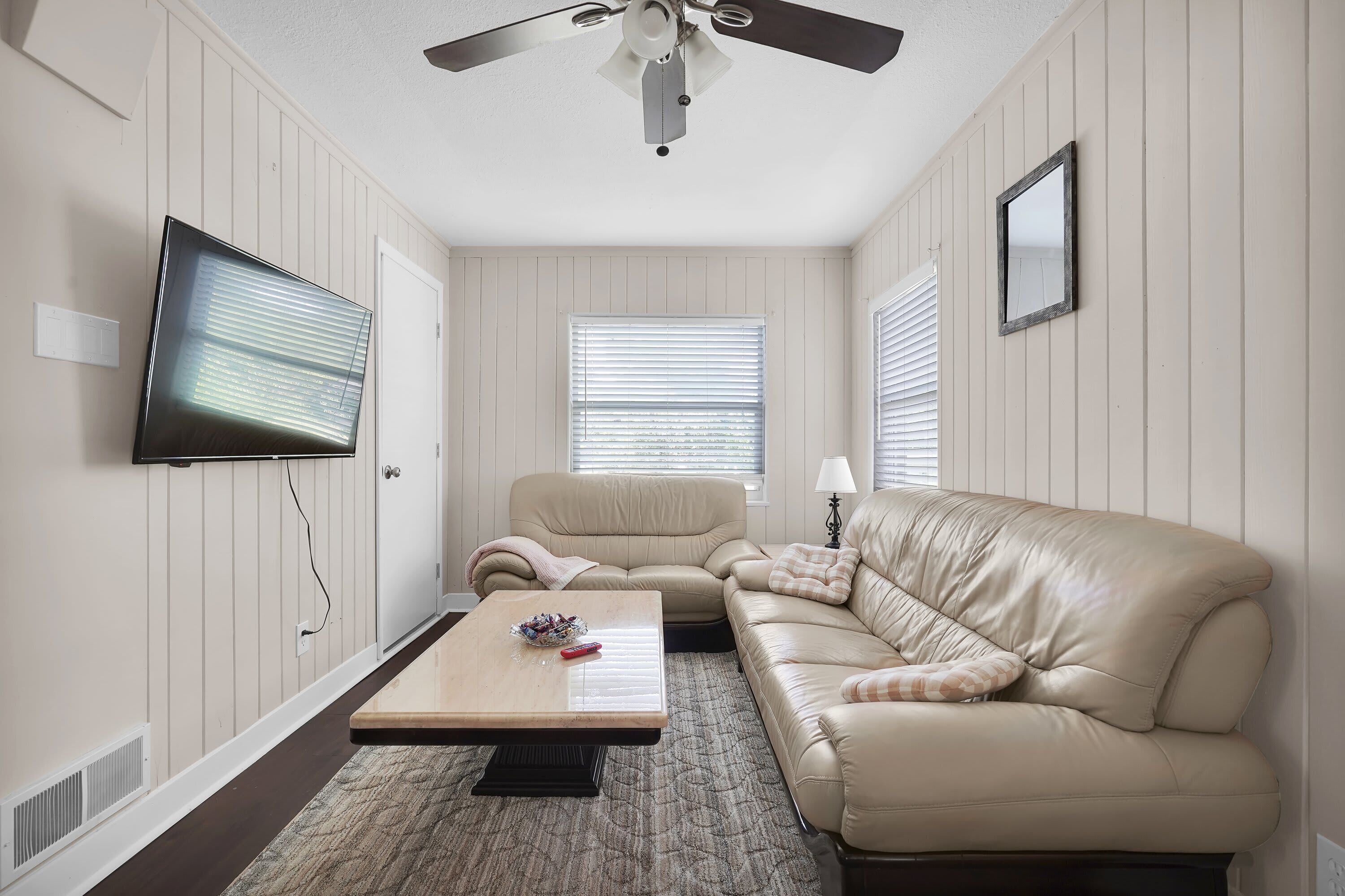 Una sala de estar con un ventilador de techo, un televisor de pantalla plana, un sofá de cuero color beige, una mesa de centro de madera y una alfombra estampada.