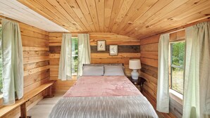 Ein Schlafzimmer einer Holzhütte mit Bett, Bank und Fenster mit Vorhängen.