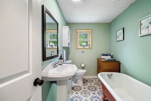 Ein Badezimmer mit einem weißen Waschbecken, einer Toilette und einer Badewanne. An der Wand hängen zwei Bilder und ein Fenster mit Blick auf Bäume.