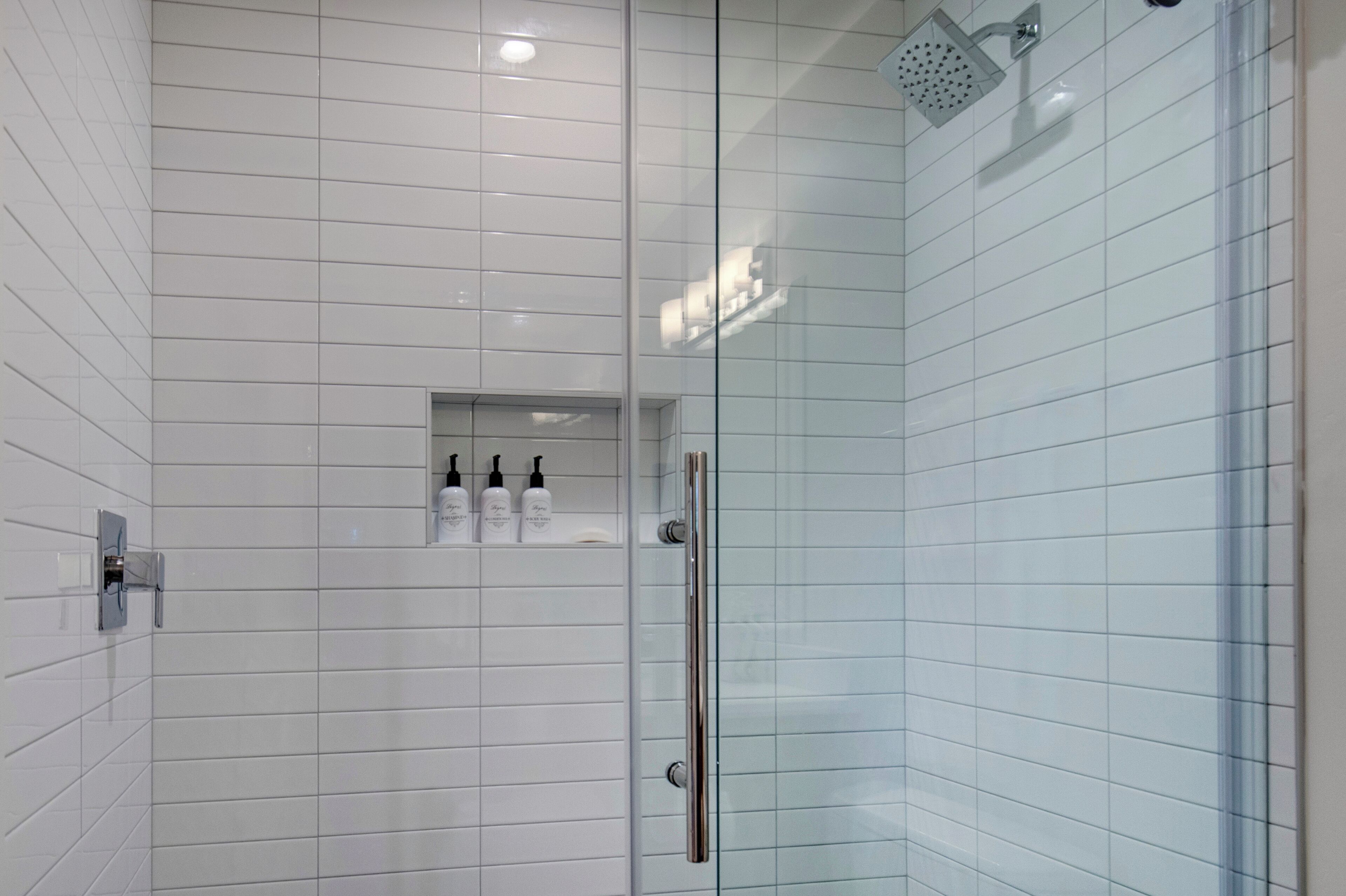 Un baño moderno con paredes de azulejos blancos, cabina de ducha de vidrio y un pequeño estante con artículos de aseo.