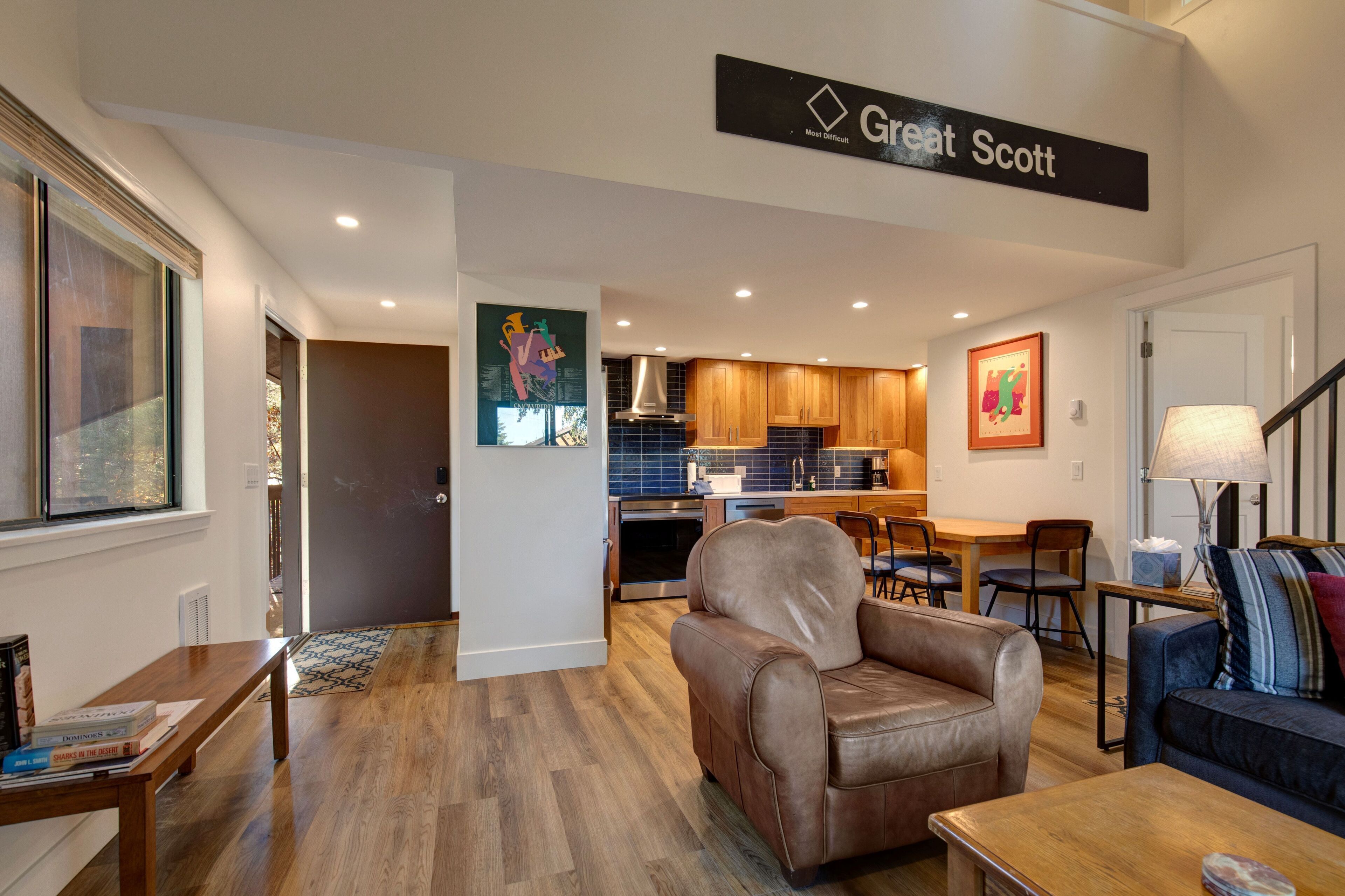 A modern living room with a leather sofa, a wooden coffee table, and a blue sofa with striped cushions. A kitchen with wooden cabinets and a dining area with a table and chairs is visible in the background.