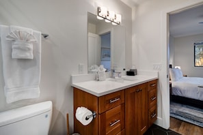 A modern bathroom with a white vanity, wooden cabinets, a large mirror with lights, a toilet, and a visible bedroom with a bed and window. - Red Pine Condos by Park City Vacations (Park City)