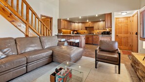 A spacious living room with a leather sofa, a glass coffee table, and a wooden chair. A kitchen with wooden cabinets and a central island is visible in the background. - Red Pine Condos by Park City Vacations (Park City)