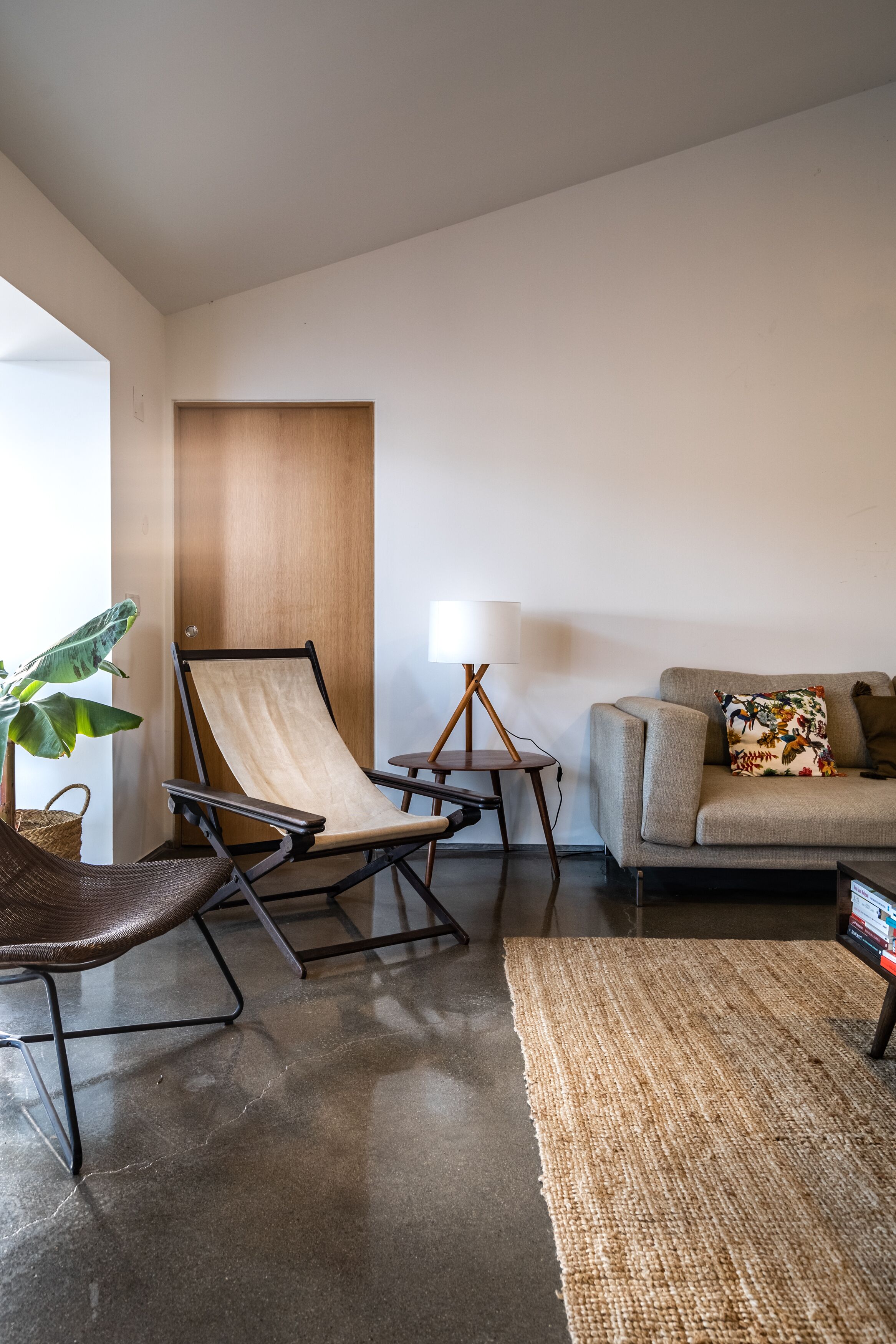 Una sala de estar moderna con suelo de concreto, un sofá color beige, una puerta de madera, un sillón, una mesita con una lámpara y una planta grande.