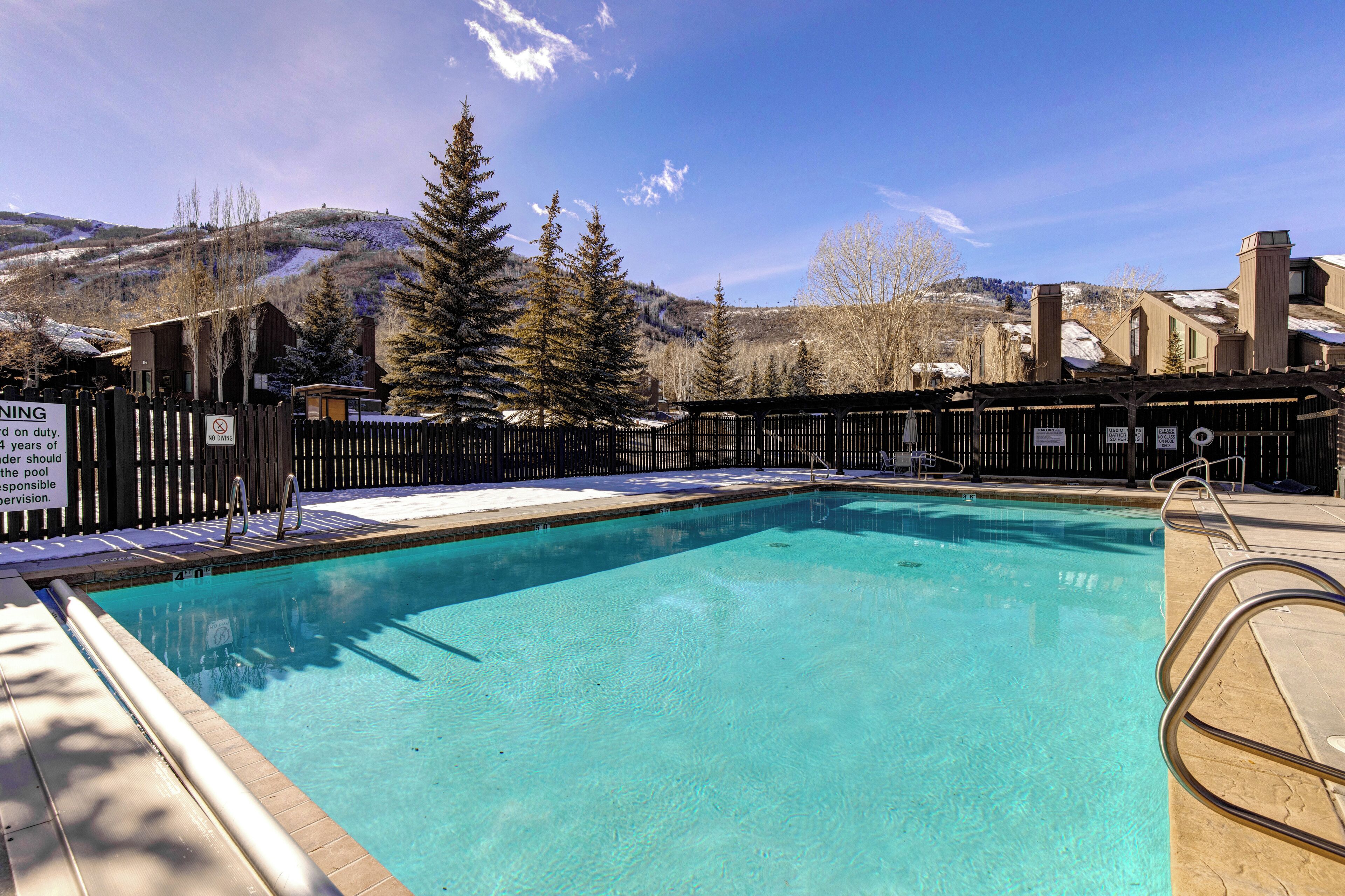 A clear swimming pool with a wooden deck, surrounded by a fence and trees, with a mountainous backdrop.