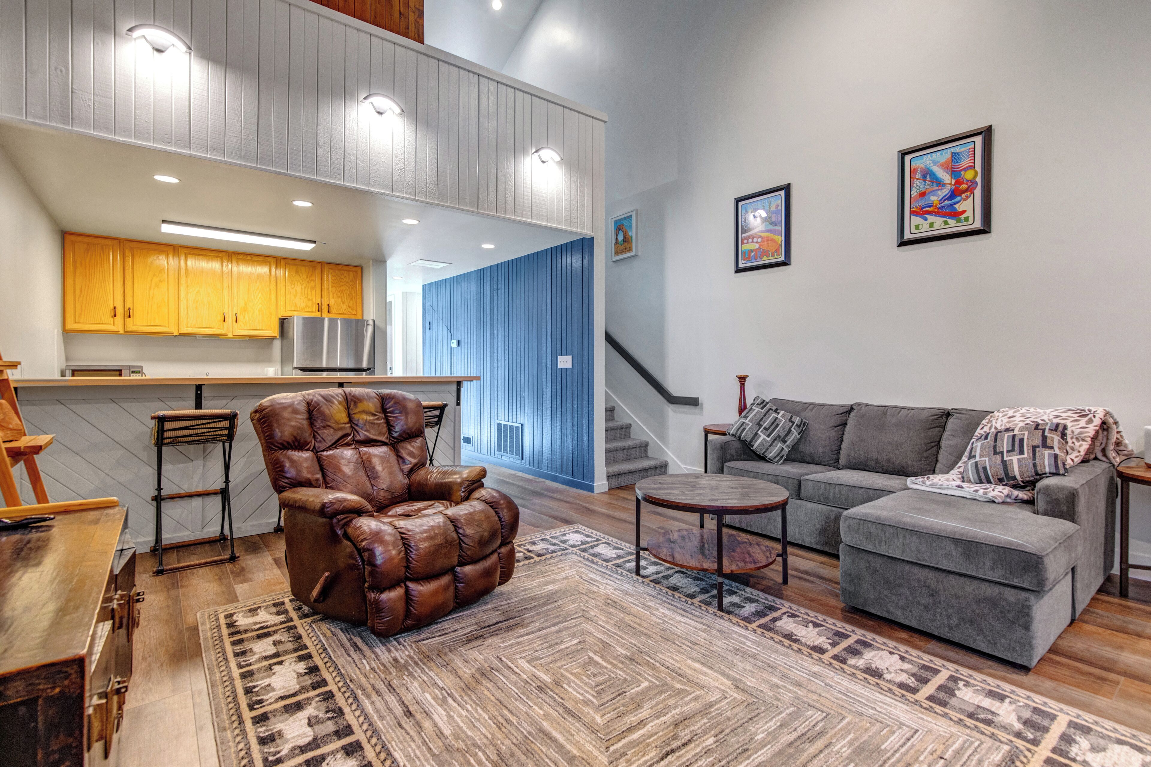 A modern living room with a leather armchair, grey sofa, and a small coffee table. A kitchen area with wooden cabinets and a stainless steel refrigerator is visible in the background.