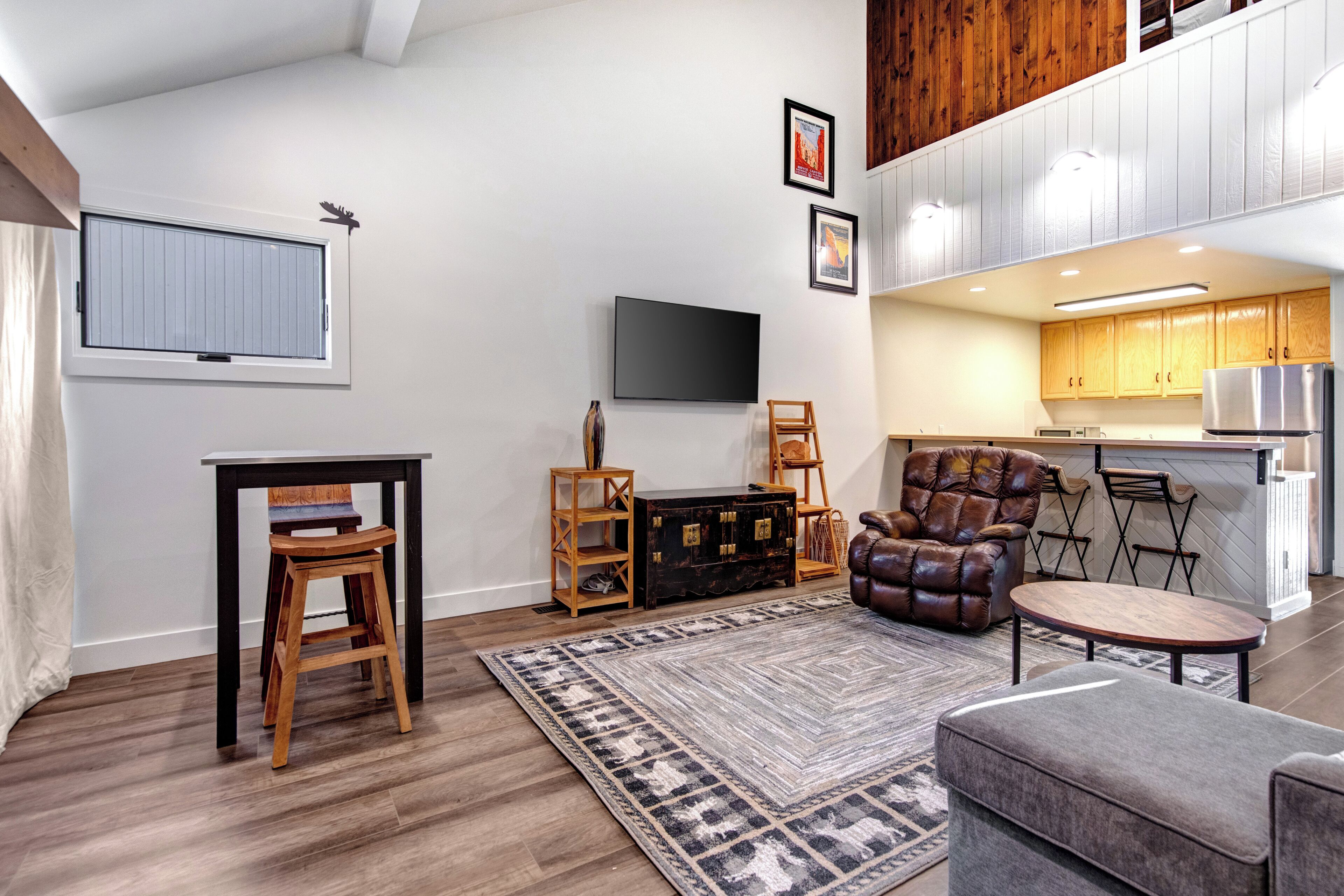 A modern living room with a flat-screen TV, a leather armchair, a dining area, and a kitchen with wooden cabinets.