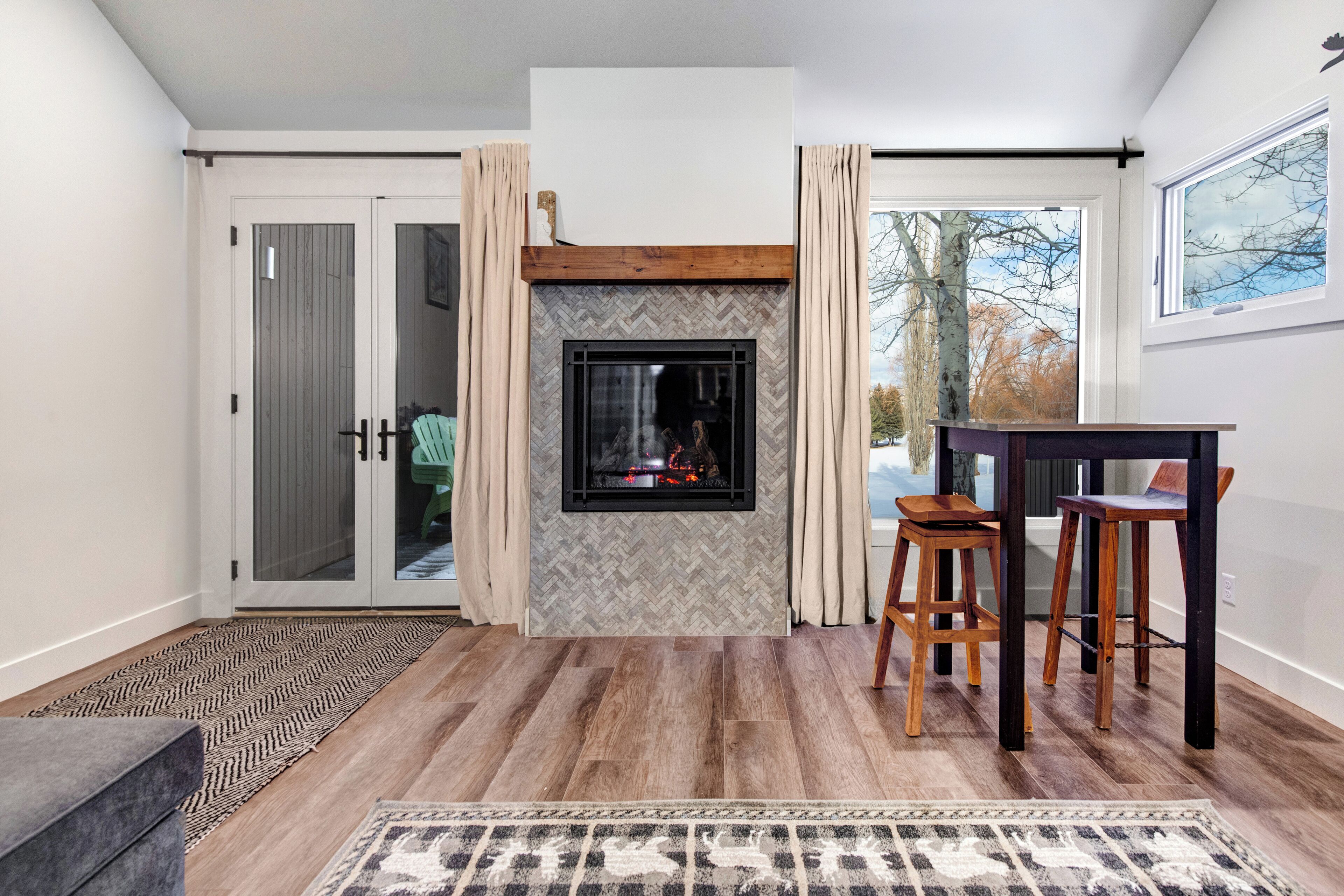 A modern living room with a fireplace, a dining area with two wooden stools, and a large window with a view of trees.