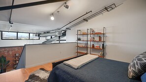 Un dormitorio estilo loft con techo alto, conductos expuestos, una estantería empotrada, una cama con cubrecama azul y vistas a la ciudad a través de amplias ventanas.