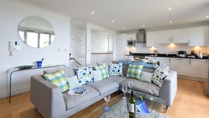 Een moderne woonkamer met een grijze bank, een glazen salontafel en een ronde spiegel. Op de achtergrond is een keuken met witte kasten en een eethoek met een tafel en stoelen te zien.
