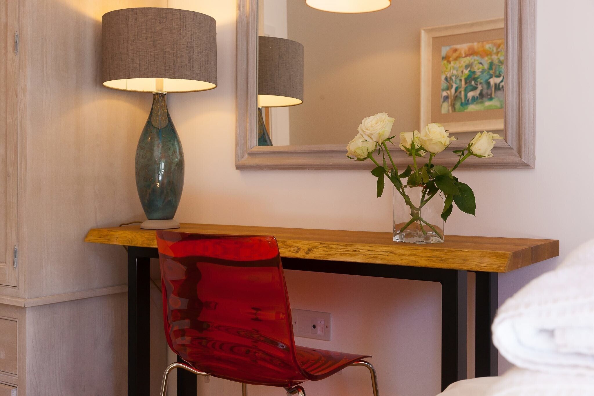 A wooden desk with a vase of white roses, a red chair, and a lamp.