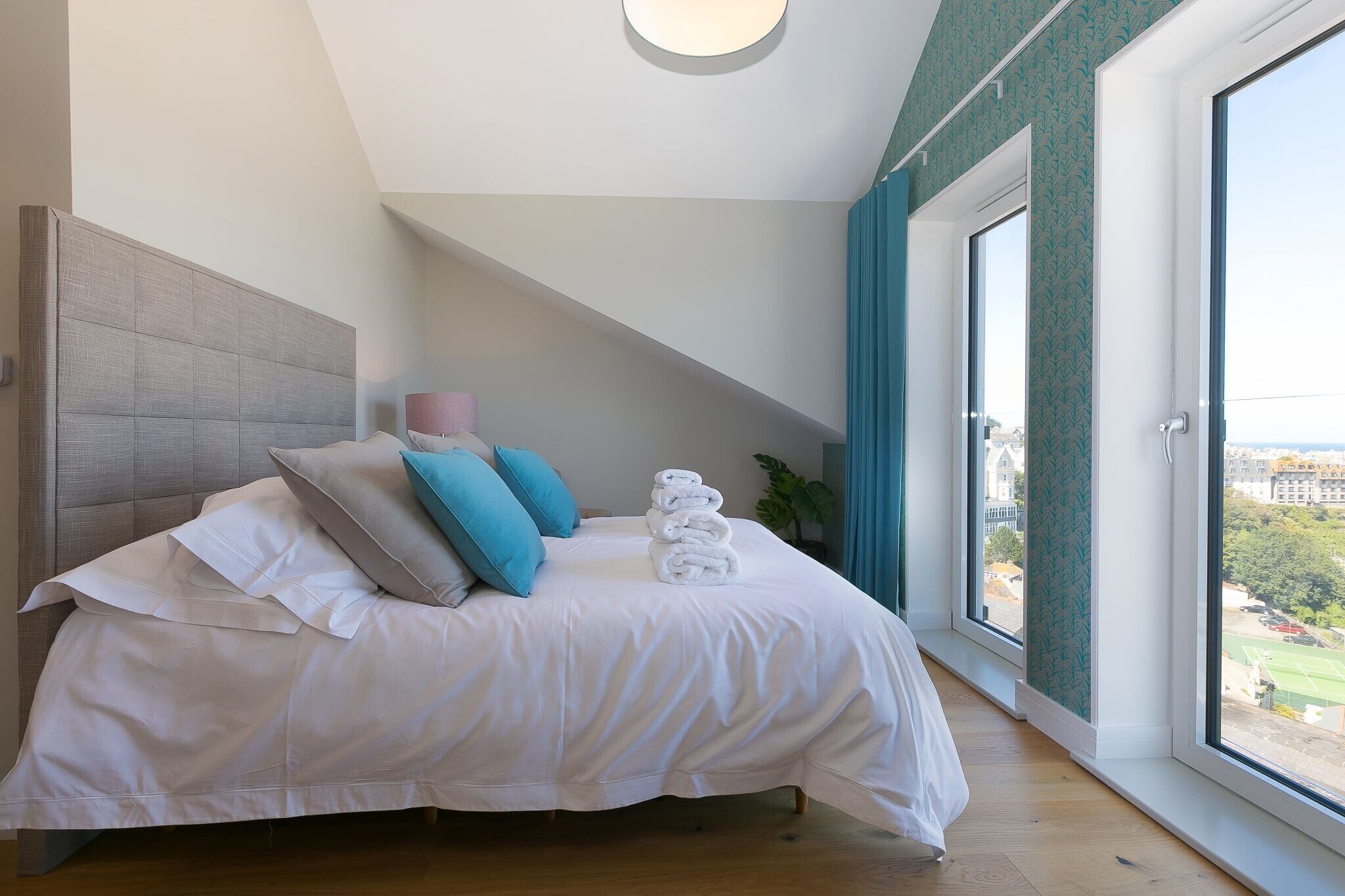 A modern bedroom with a large bed, a headboard, a potted plant, and a view of the city through large windows.