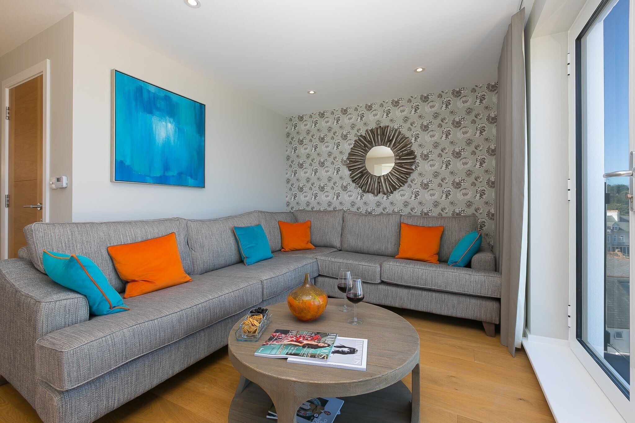 A modern living room with a grey sectional sofa, orange and blue cushions, a coffee table with magazines and a vase, and a large abstract painting on the wall.