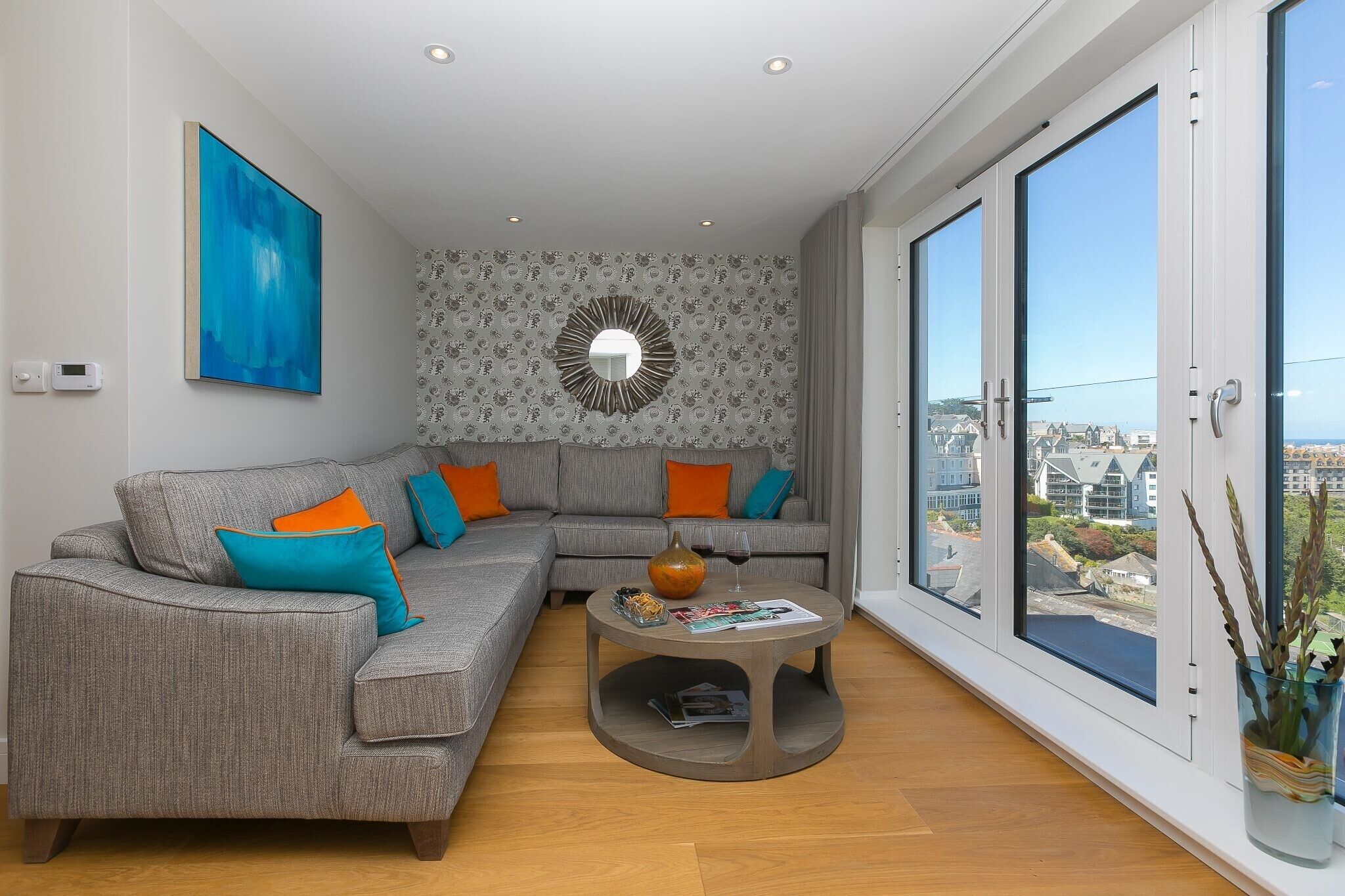 Ein modernes Wohnzimmer mit einer grauen Eckcouch, einem runden Couchtisch und einem großen Fenster mit Blick auf die Stadt.
