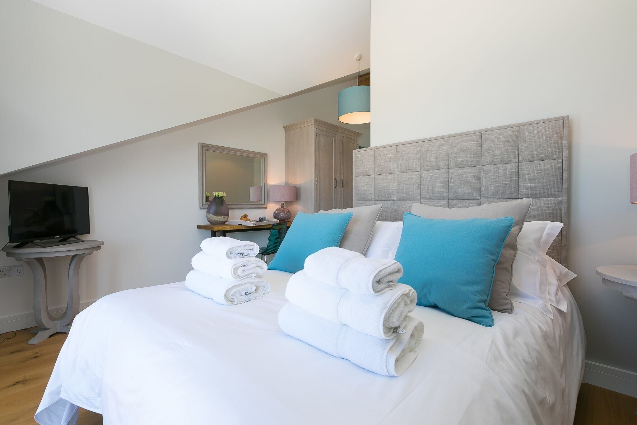 Una cama bien hecha con almohadas blancas y azules, un televisor sobre una mesita y un espejo en la pared.