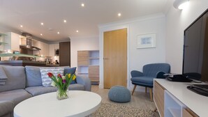 A modern living room with a sofa, coffee table, and a blue chair. There is a kitchen in the background with a microwave and cabinets. - The Sanctuary (St Ives)