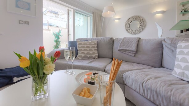A modern living room with a grey sofa, a white table with a vase of flowers, and a plate of sushi. - The Sanctuary (St Ives)