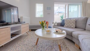 A modern living room with a flat-screen TV, a white coffee table with flowers, a grey sofa, and a large window with blinds. - The Sanctuary (St Ives)