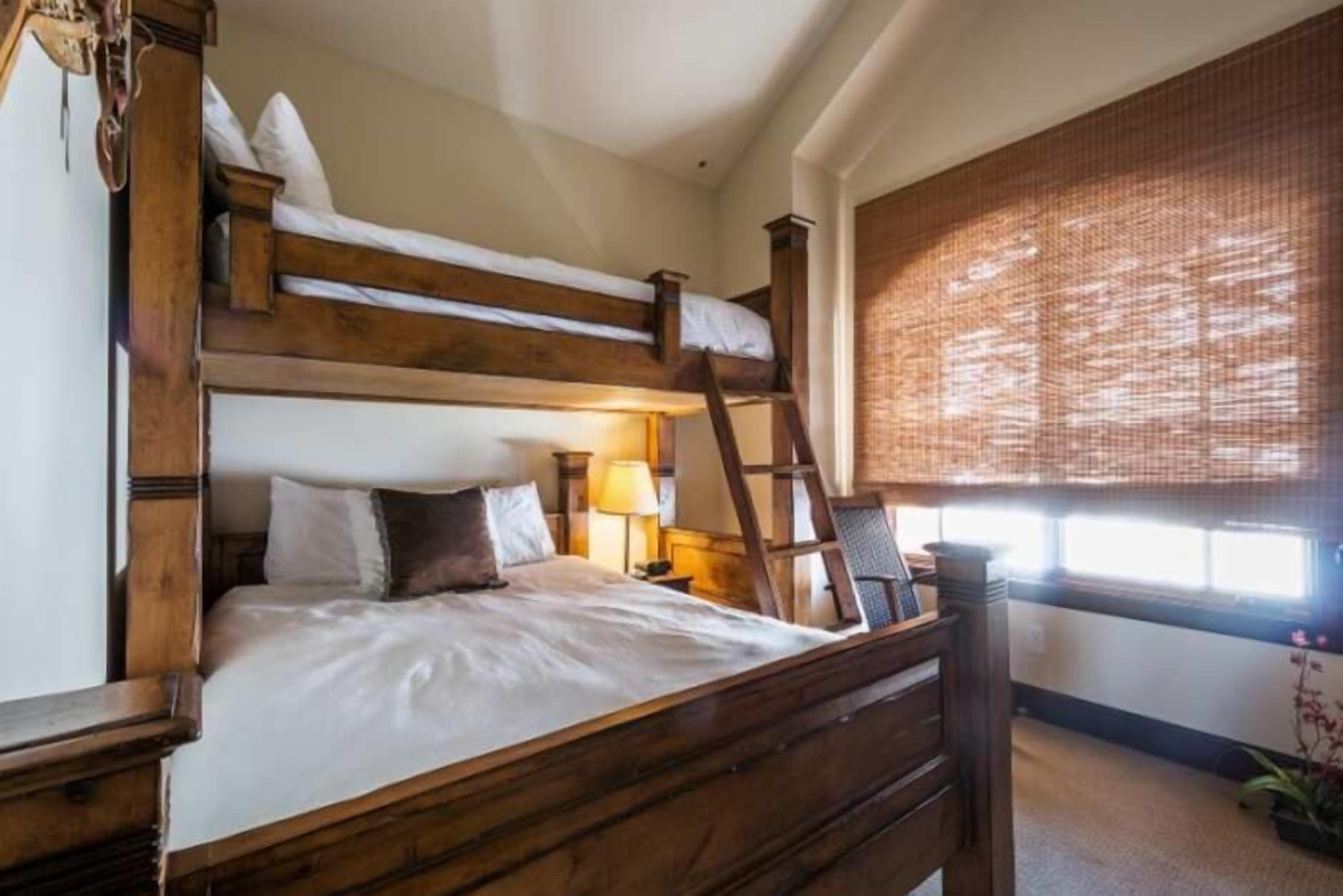 Un lit superposé avec une échelle, une table de chevet, une lampe et une fenêtre avec un store en bambou.