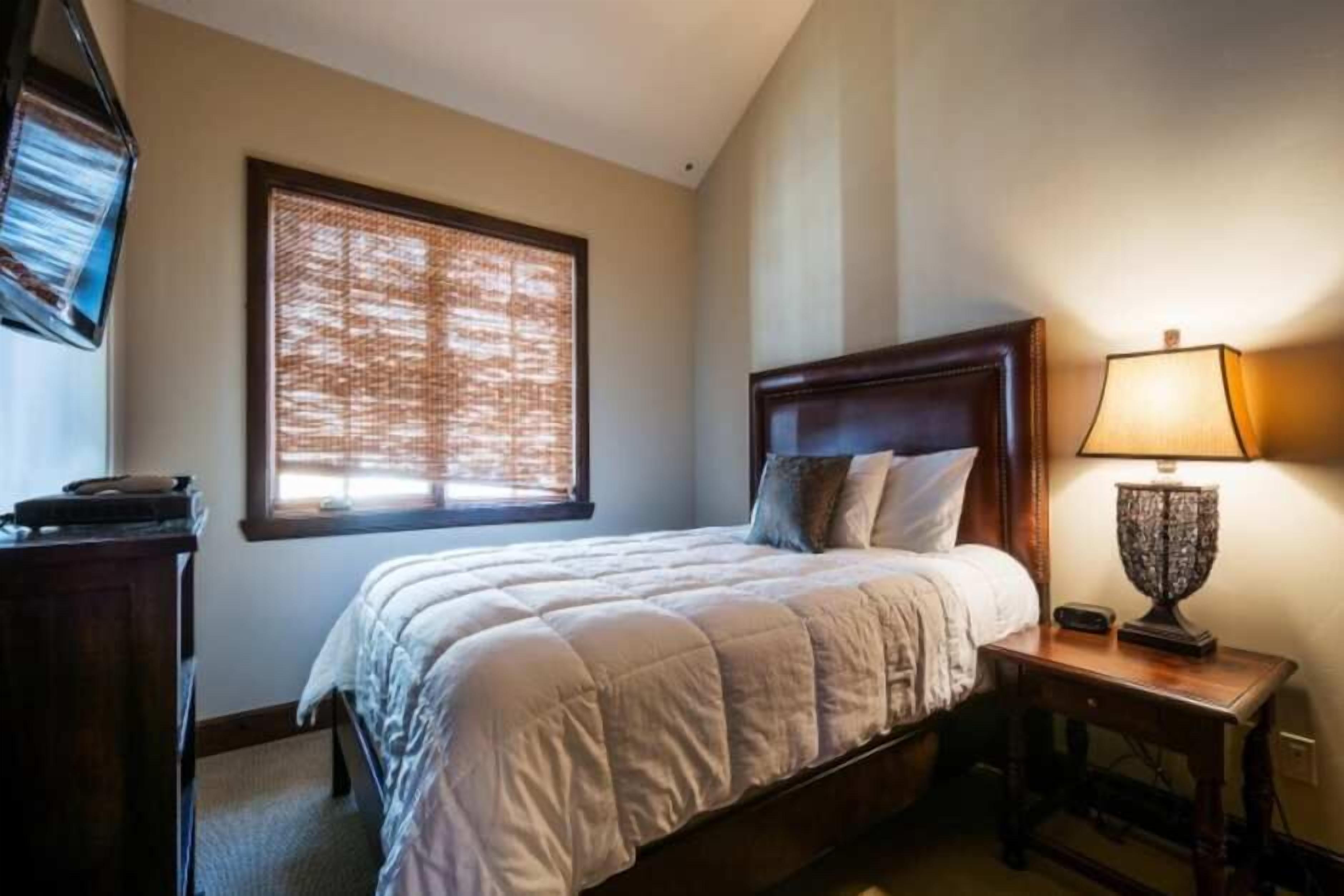 A bedroom with a bed, a nightstand with a lamp, a window with blinds, and a television on the wall.