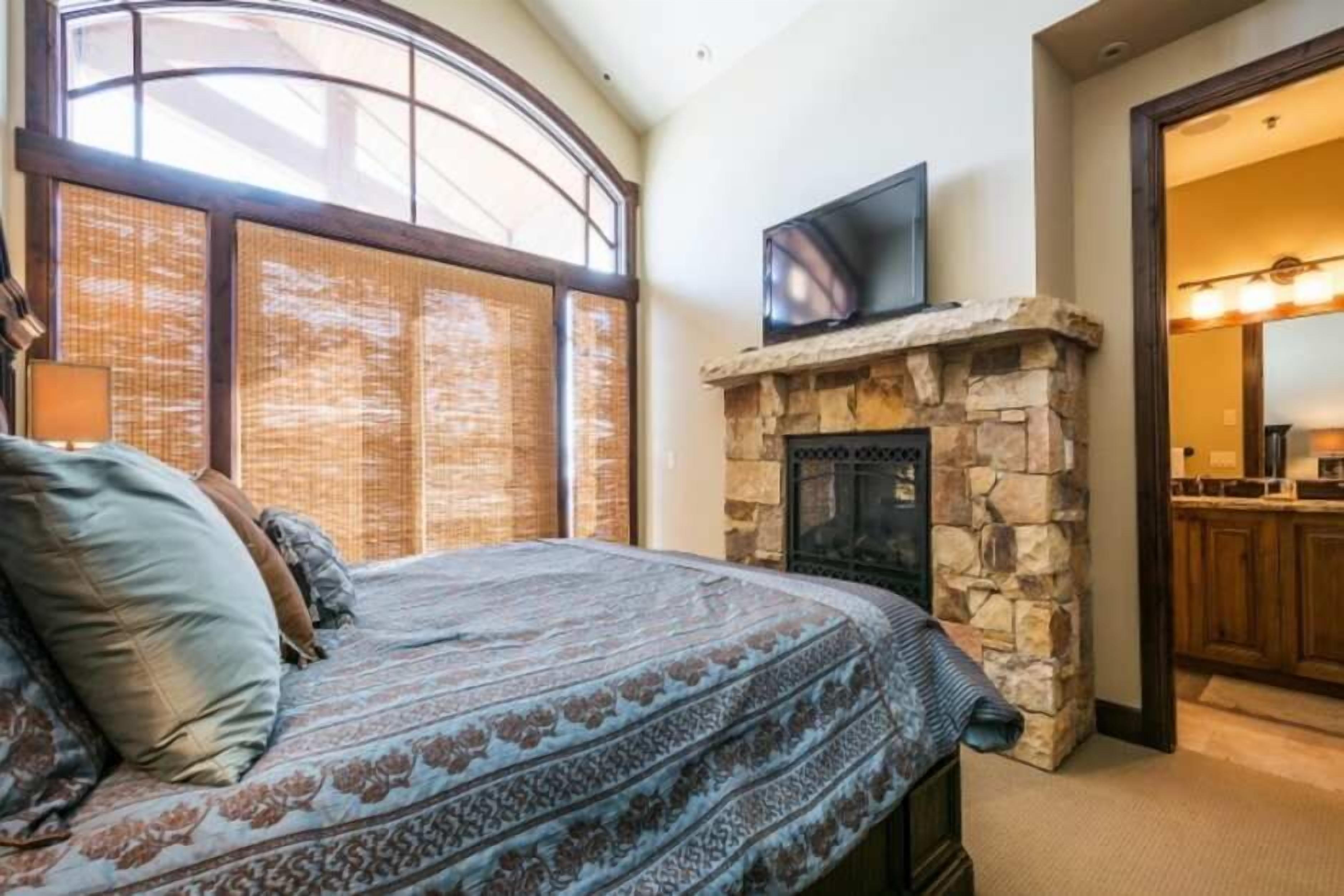 Un dormitorio con una cama grande, una chimenea de piedra, un televisor colocado encima y vistas de la cocina a través de una puerta abierta.