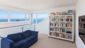 A room with a blue sofa, a white bookshelf, and a view of the sea through large windows. - Shamal (St Ives)